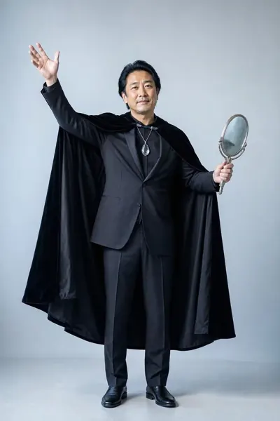 Illusionist, ein Mann mittleren Alters in schwarzem Anzug und Umhang hält einen Handspiegel und hebt einen Arm.